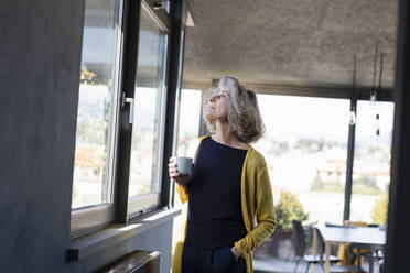 Nachdenkliche Frau mit Kaffeetasse, die zu Hause am Fenster steht - EIF00438