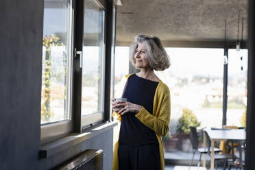 Lächelnde Frau mit Kaffeetasse, die durch das Fenster schaut, während sie zu Hause steht - EIF00439