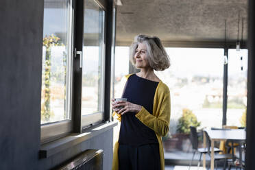 Smiling woman with coffee cup looking through window while standing at home - EIF00439