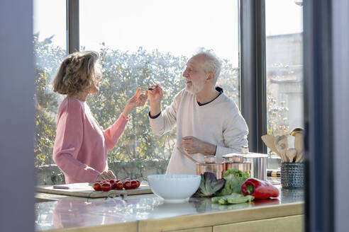 Smiling senior man feeding woman while standing at home - EIF00472