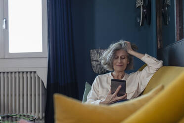 Senior man with hand in hair using mobile phone while sitting on sofa at home - EIF00495