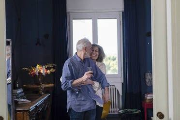 Lächelnde Frau umarmt Mann, der ein Weinglas hält, während sie zu Hause steht - EIF00515