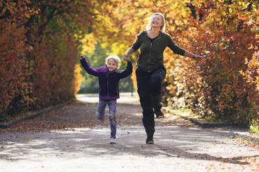Mother and daughter holding hands and running in park during autumn - PSIF00448