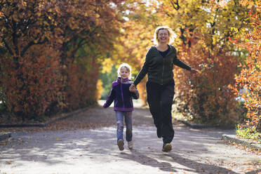 Happy mother and daughter holding hands and running in park during autumn - PSIF00457