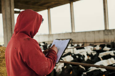 Farmer writing on clipboard while standing at dairy farm - ACPF01179