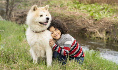 Smiling boy leaning on dog while sitting on grass in nature - JCCMF01345