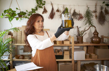 Frau hält Becher mit Olivenöl bei der Herstellung von Seife in einer Werkstatt - VEGF04017