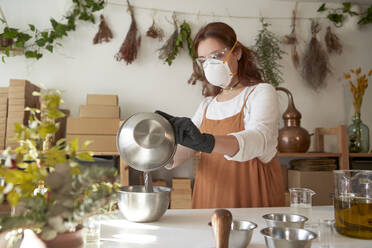 Woman with protective eyewear and mask mixing ingredient in bowls while making soap in workshop - VEGF04025