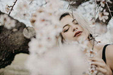 Young woman with eyes closed standing under blossoming almond tree - GMLF01026