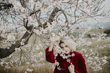 Smiling young woman in red winter coat standing under blossoming almond tree - GMLF01030