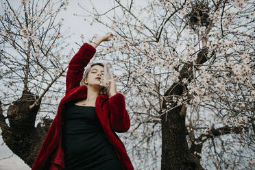 Junge Frau in rotem Wintermantel steht im Frühling unter einem blühenden Mandelbaum - GMLF01033