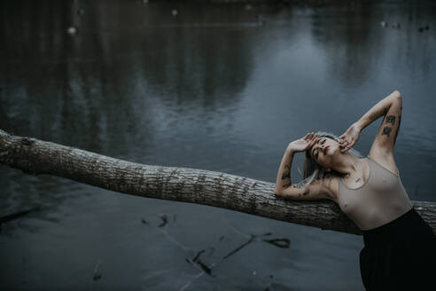 Young woman with eyes closed leaning on fallen tree against lake - GMLF01044