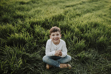 Fröhlicher Junge mit gefalteten Händen auf einer Wiese sitzend - GMLF01057