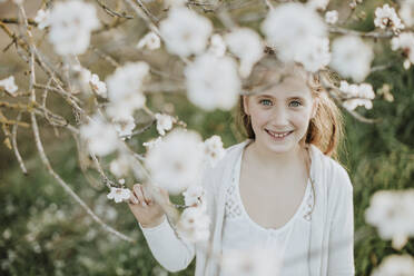 Nettes Mädchen lächelt, während es einen Zweig einer Mandelbaumblüte im Frühling hält - GMLF01064