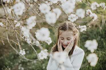 Innocent girl with hands on cheeks standing by almond blossoms - GMLF01065