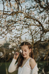 Nettes Mädchen bedeckt Auge von Zweig der Mandelbaum Blume im Frühling - GMLF01068