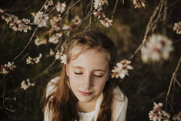 Girl standing with eyes closed under branches of almond tree during springtime - GMLF01071