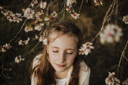 Mädchen steht mit geschlossenen Augen unter den Zweigen eines Mandelbaums im Frühling - GMLF01071