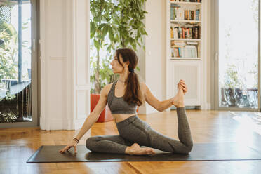 Flexible Frau, die ihre Beine streckt, während sie zu Hause Yoga übt - GMCF00073