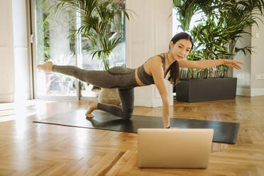 Beautiful yoga instructor practicing bird dog pose during online class through laptop - GMCF00086