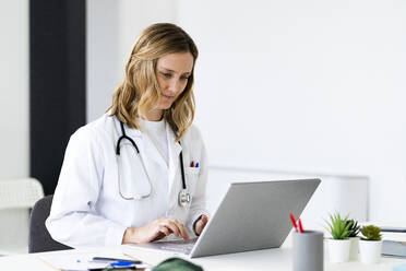 Mittlere erwachsene Ärztin, die einen Laptop benutzt, während sie am Schreibtisch in einer medizinischen Klinik sitzt - GIOF11605