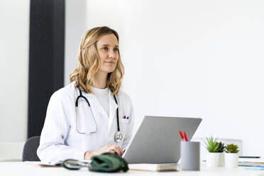 Blond female doctor sitting with laptop at desk looking away - GIOF11606