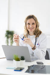 Smiling female doctor sitting with laptop at desk in medical clinic - GIOF11611
