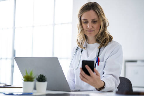 Blonde Ärztin, die ein Mobiltelefon benutzt, sitzt am Schreibtisch in einer medizinischen Klinik - GIOF11612