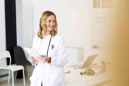 Lächelnde Mitarbeiterin des Gesundheitswesens mit Tablet, die sich in einer medizinischen Klinik auf den Schreibtisch stützt - GIOF11624
