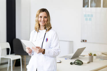 Mittlere erwachsene Ärztin mit digitalem Tablet, die sich im Stehen auf den Schreibtisch stützt - GIOF11625