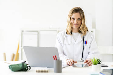 Blond female medical professional sitting with tablet and laptop at desk - GIOF11626