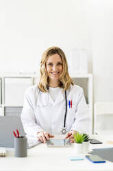 Smiling female doctor sitting with tablet and laptop at desk - GIOF11627