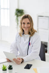 Smiling female doctor sitting with tablet and smart phone at desk - GIOF11629