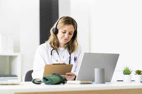 Medizinische Mitarbeiterin schreibt auf einem Klemmbrett, während sie eine Online-Konsultation über einen Laptop in einer medizinischen Klinik durchführt - GIOF11637