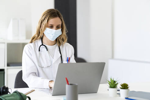 Ärztin mit Schutzmaske, die am Schreibtisch sitzend am Laptop arbeitet - GIOF11640