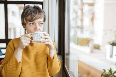 Reife Frau trinkt Kaffee und schaut durch das Fenster zu Hause - LJF02091