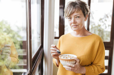 Short hair woman having breakfast while standing at glass window - LJF02092