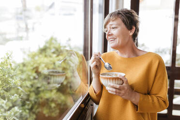 Happy mature woman having breakfast while looking through window at home - LJF02093