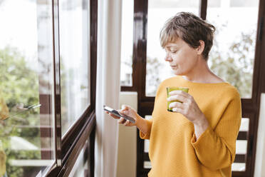 Reife Frau mit Trinkglas, die ihr Smartphone benutzt, während sie am Glasfenster steht - LJF02094