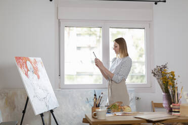 Weibliche Künstlerin, die ein Foto von einem Gemälde mit ihrem Smartphone macht, während sie im Heimstudio steht - EIF00534