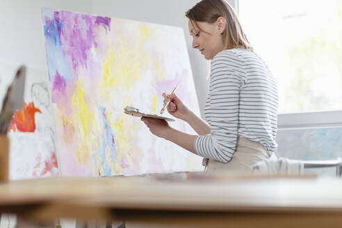 Junge Frau hält Farbpalette beim Malen abstrakter Leinwand im Heimstudio - EIF00536
