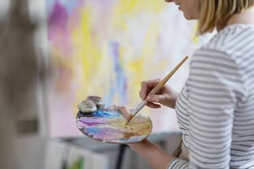 Female artist mixing colors on palette while painting abstract canvas at home studio - EIF00538