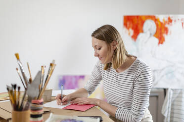 Female artist writing in book while sitting at table in home studio - EIF00540