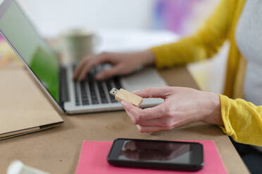 Woman holding USB stick by laptop and mobile phone on table at home - EIF00547