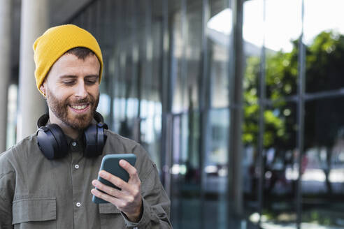 Lächelnder gut aussehender Mann mit Kopfhörern, der in der Nähe eines Gebäudes ein Smartphone benutzt - PNAF00907