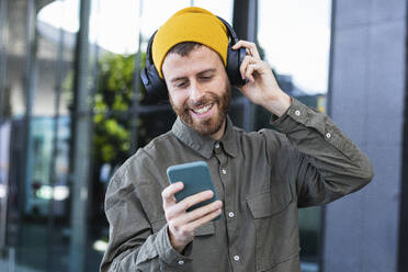 Man listening music on headphones through mobile phone near building - PNAF00908