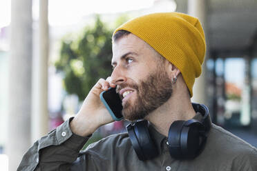 Fashionable man with headphones talking on smart phone while looking away at footpath - PNAF00911