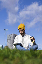 Mittlerer erwachsener Mann, der einen Kaffee trinkt, während er ein digitales Tablet im Park gegen den Himmel an einem sonnigen Tag benutzt - PNAF00914