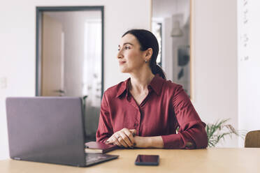 Smiling businesswoman with laptop looking away while sitting at table in home office - KMKF01574