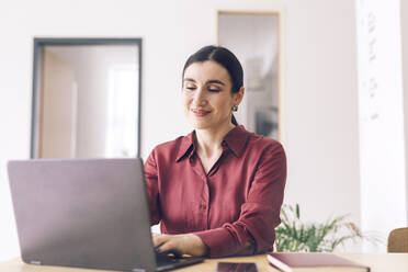 Lächelnde Unternehmerin, die einen Laptop benutzt, während sie am Tisch im Heimbüro sitzt - KMKF01575